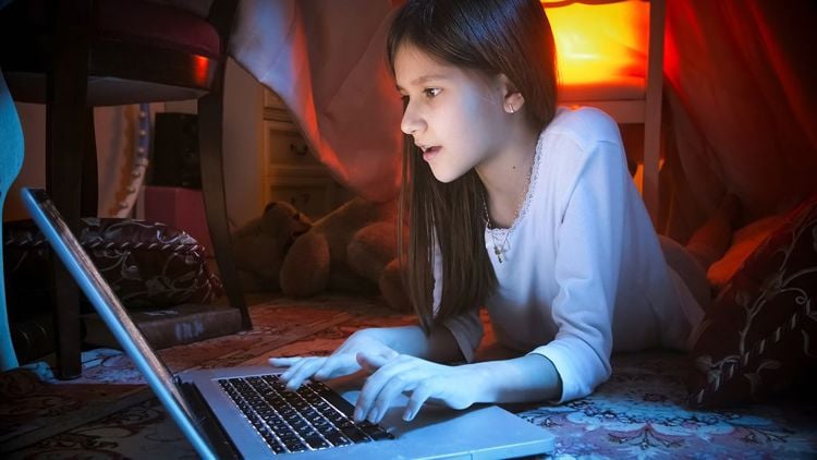 Mørkhåret jente med hvit genser ligger under et bord i halvmørket og spiller på en pc