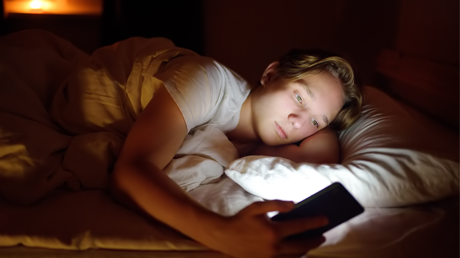 En gutt ligger i senga og ser på mobilen sin
