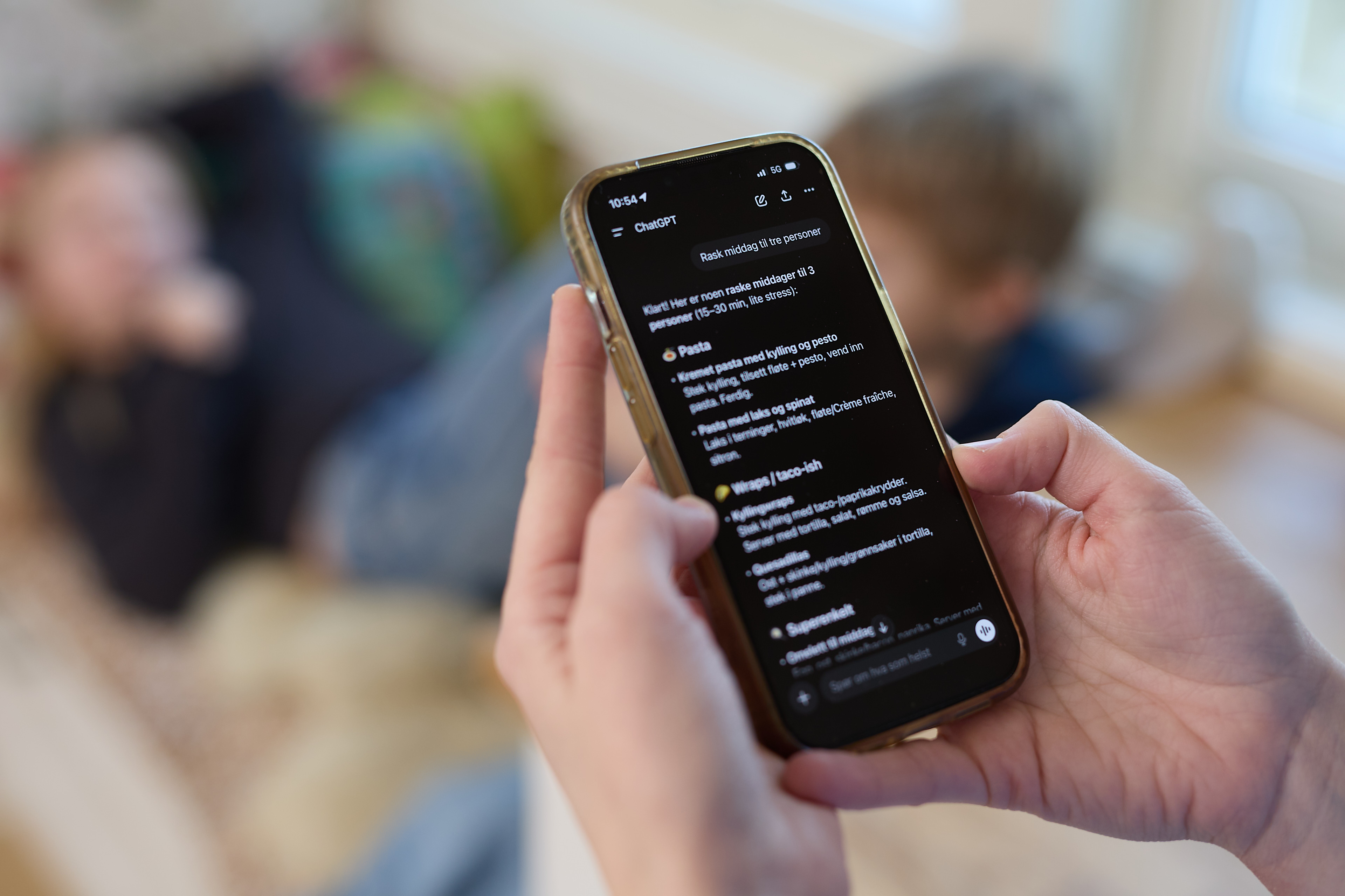 En person holder en mobiltelefon og leser en samtale med ChatGPT p&aring; skjermen. I bakgrunnen, ute av fokus, sitter to personer i en sofa i en stue.