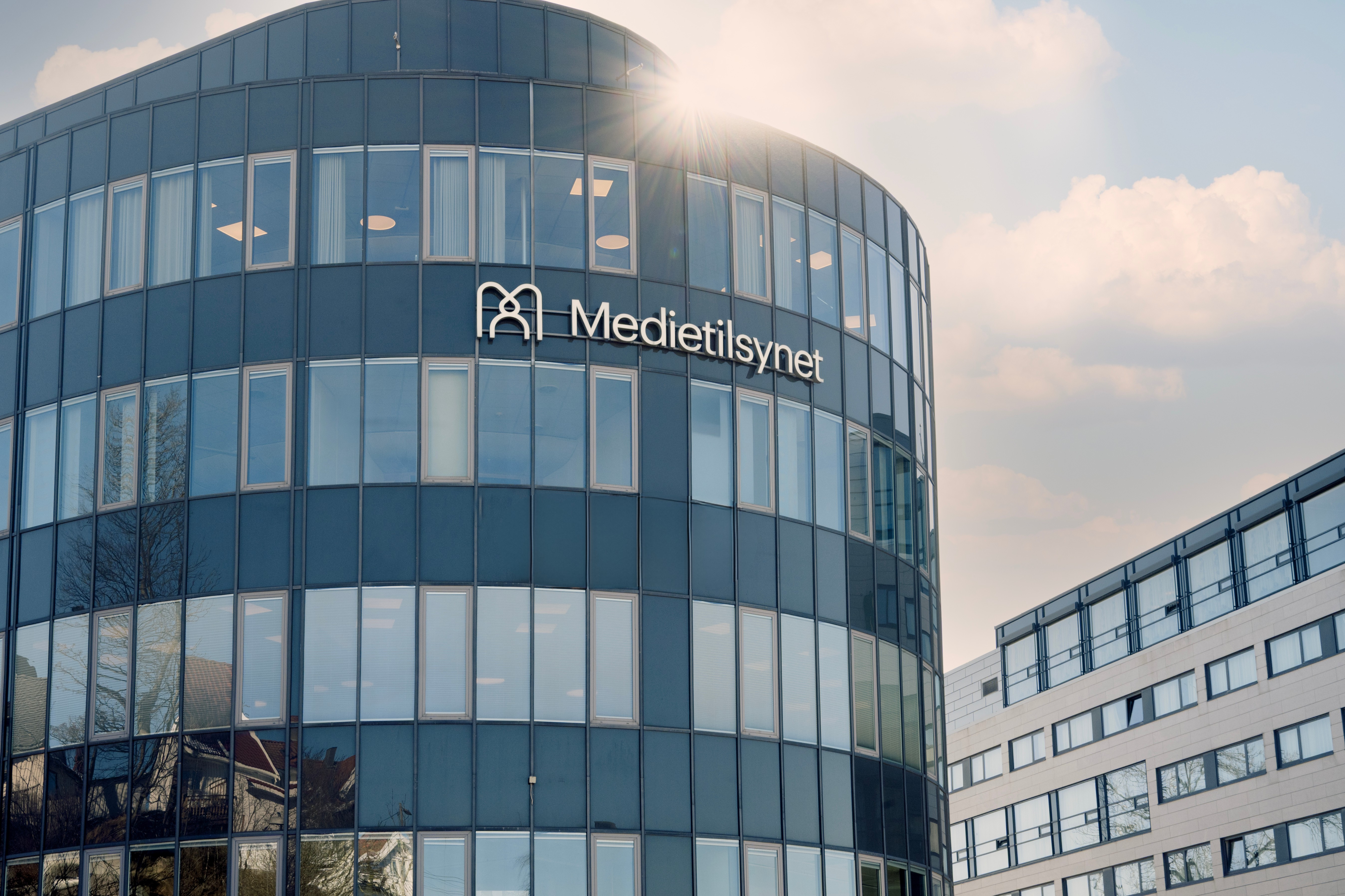 Moderne kontorbygg med buet glassfasade og skiltet &laquo;Medietilsynet&raquo;, sett i dagslys med sol som skinner over bygget.