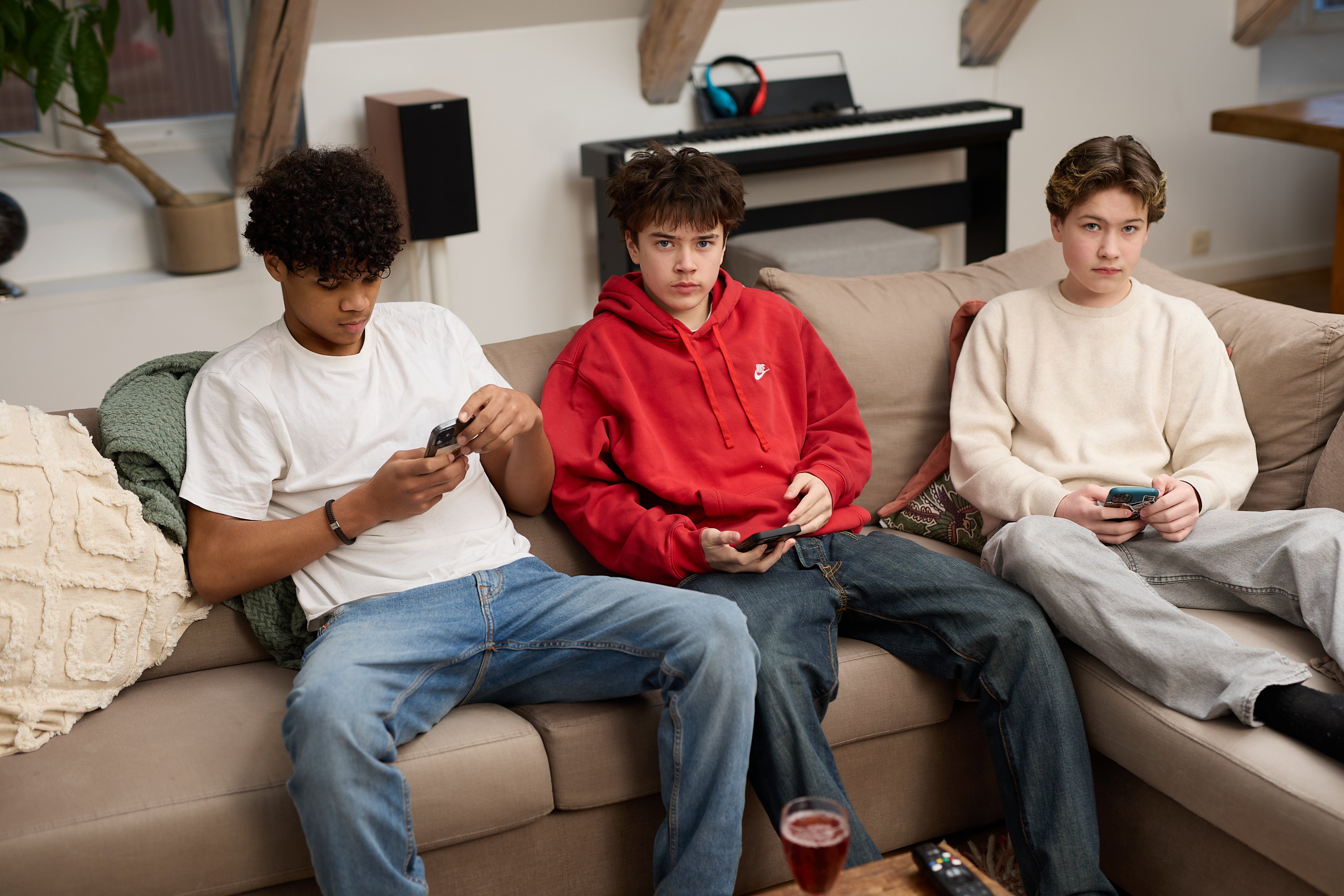 Tre ten&aring;ringsgutter sitter i en beige sofa i en stue og ser p&aring; hver sin mobiltelefon. Gutten til venstre har hvit t-skjorte og jeans, gutten i midten har r&oslash;d hettegenser, og gutten til h&oslash;yre har lys genser og gr&aring; bukse. I bakgrunnen st&aring;r et elektrisk piano med hodetelefoner p&aring;, og det ligger en fjernkontroll og et glass med r&oslash;d drikke p&aring; bordet foran dem.