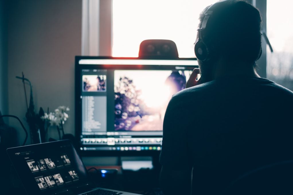 Mann fotografert bakfra mens han sitter ved arbeidsplassen og redigerer på en skjerm foran seg.