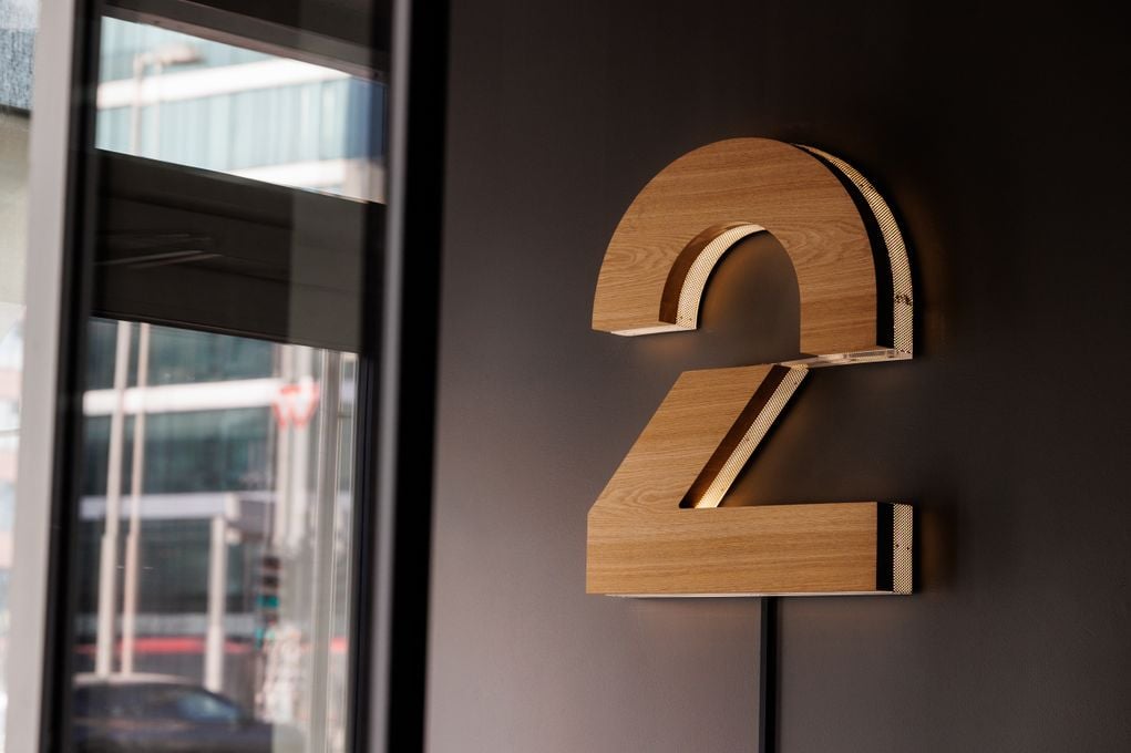 TV 2-logo på innendørs vegg. To-tallet i eik med opplyst bakgrunn mot mørkt malt vegg.