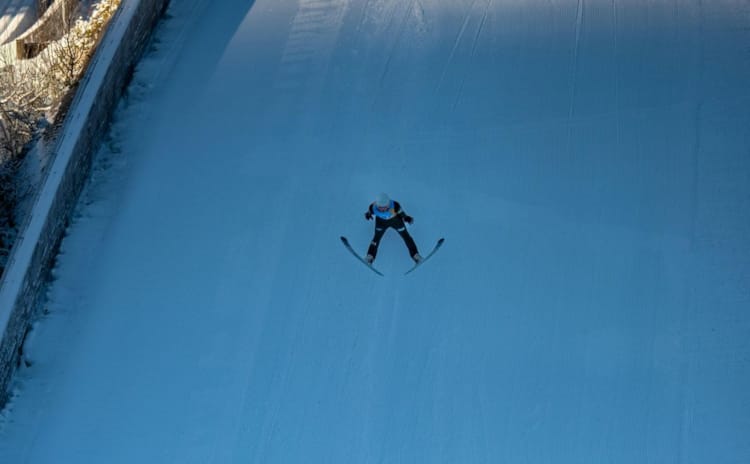 En skihopper svever gjennom luften over en hoppbakke, sett ovenfra, med skiene i V-formasjon. Bakken under er preparert og dekket av snø. Bildet gir et inntrykk av fart, høyde og presisjon.