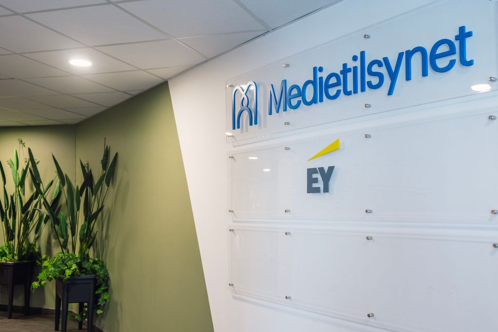 Bilde fra inngangspartiet til Medietilsynets kontor. Det er grønn vegg og to grønne planter, og på et hvitt felt på veggen står Medietilsynets logo øverst.