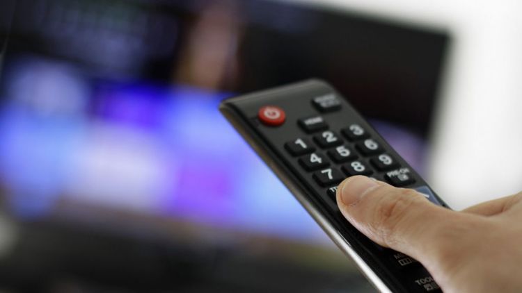Nærbilde av fjernkontroll og en tommel. Uskarpt i bakgrunnen skimter vi en tv-skjerm.