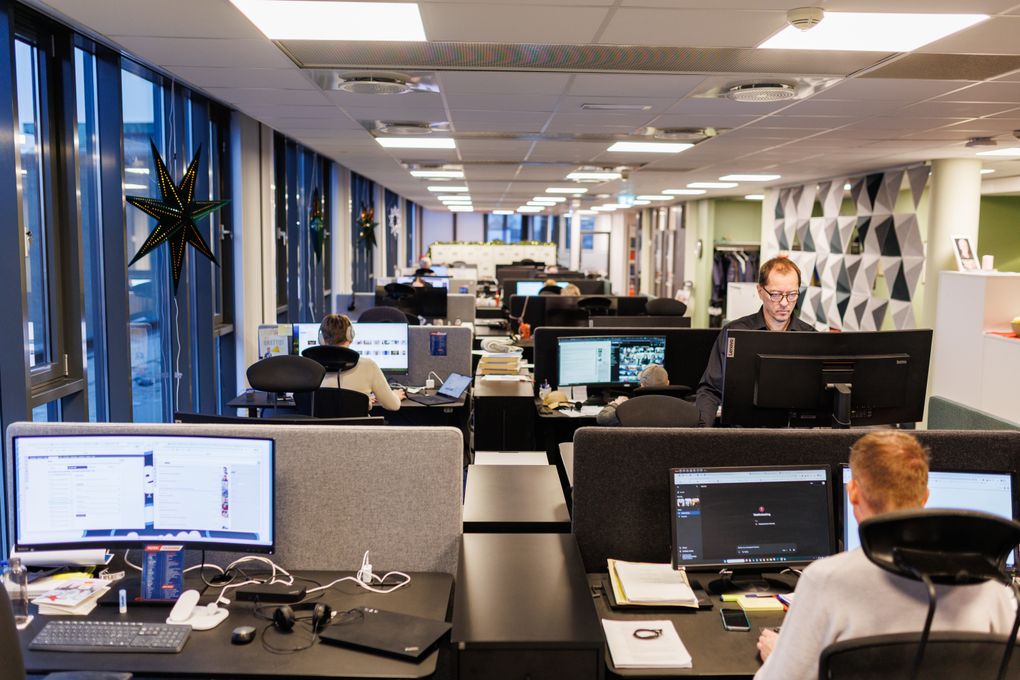 Oversiktsbillete frå avisredaksjon. Stort, lyst, åpent landskap med fire og fire arbeidsplassar ved siden av og mot kvarandre. Ein person står og jobbar med ansikt mot kamera, mens vi ser fleire bakhoder framfor opplyste pc-skjermar.