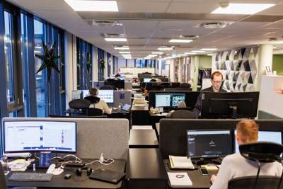 Oversiktsbillete frå avisredaksjon. Stort, lyst, åpent landskap med fire og fire arbeidsplassar ved siden av og mot kvarandre. Ein person står og jobbar med ansikt mot kamera, mens vi ser fleire bakhoder framfor opplyste pc-skjermar.