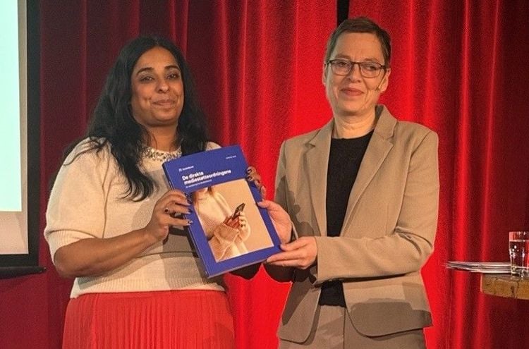 Kultur- og likestillingsminister Lubna Jaffery, og direktør i Medietilsynet, Mari Velsand, står foran et rødt sceneteppe og holder sammen rapporten ‘De direkte mediestøtteordningene’
