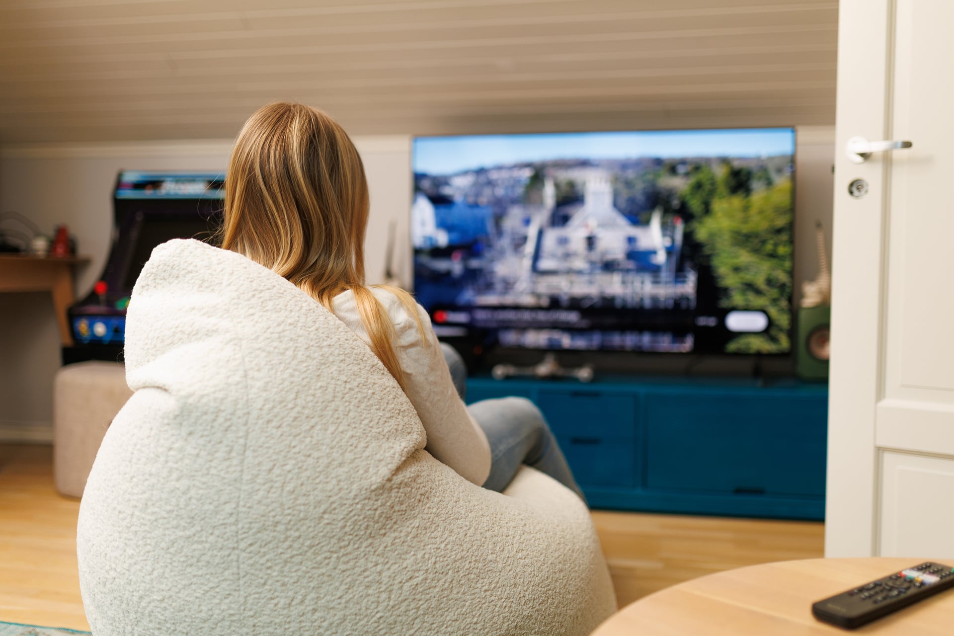 A child sits in a soft chair and watches television in a living room, with a remote control on a table in the foreground.