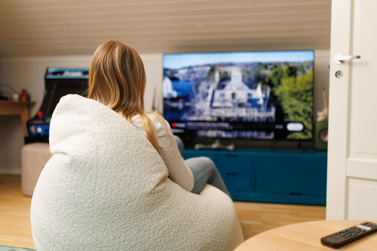 A child sits in a soft chair and watches television in a living room, with a remote control on a table in the foreground.
