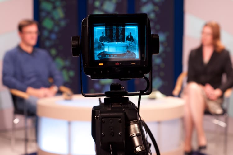 Vi ser baksiden av et kamera som filmer to personer sittende i et tv-studio.