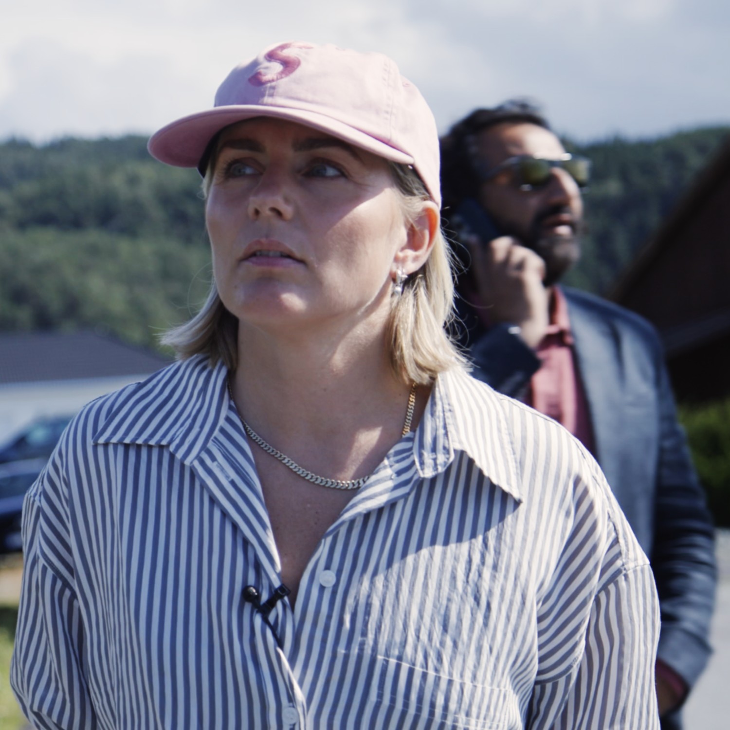To personer st&aring;r utend&oslash;rs i sollys, med skog og bygninger i bakgrunnen. I forgrunnen st&aring;r en kvinne med rosa caps og stripete skjorte og ser alvorlig til siden. Bak henne st&aring;r en mann i dressjakke og solbriller med en telefon mot &oslash;ret. Bildet gir inntrykk av en reportasje- eller dokumentarscene.