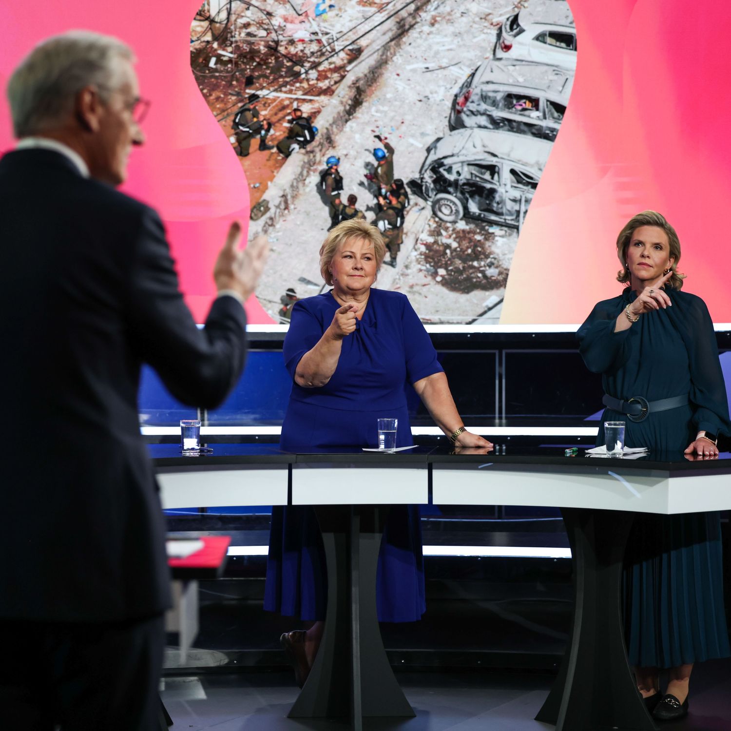 TV-studio under en politisk partilederdebatt. En mann st&aring;r med ryggen delvis mot kamera og henvender seg til to kvinnelige politikere bak hvert sitt podium. Begge politikerne gestikulerer med hendene. P&aring; skjermen bak dem vises et nyhetsbilde av &oslash;delagte biler og personer p&aring; et skadested.