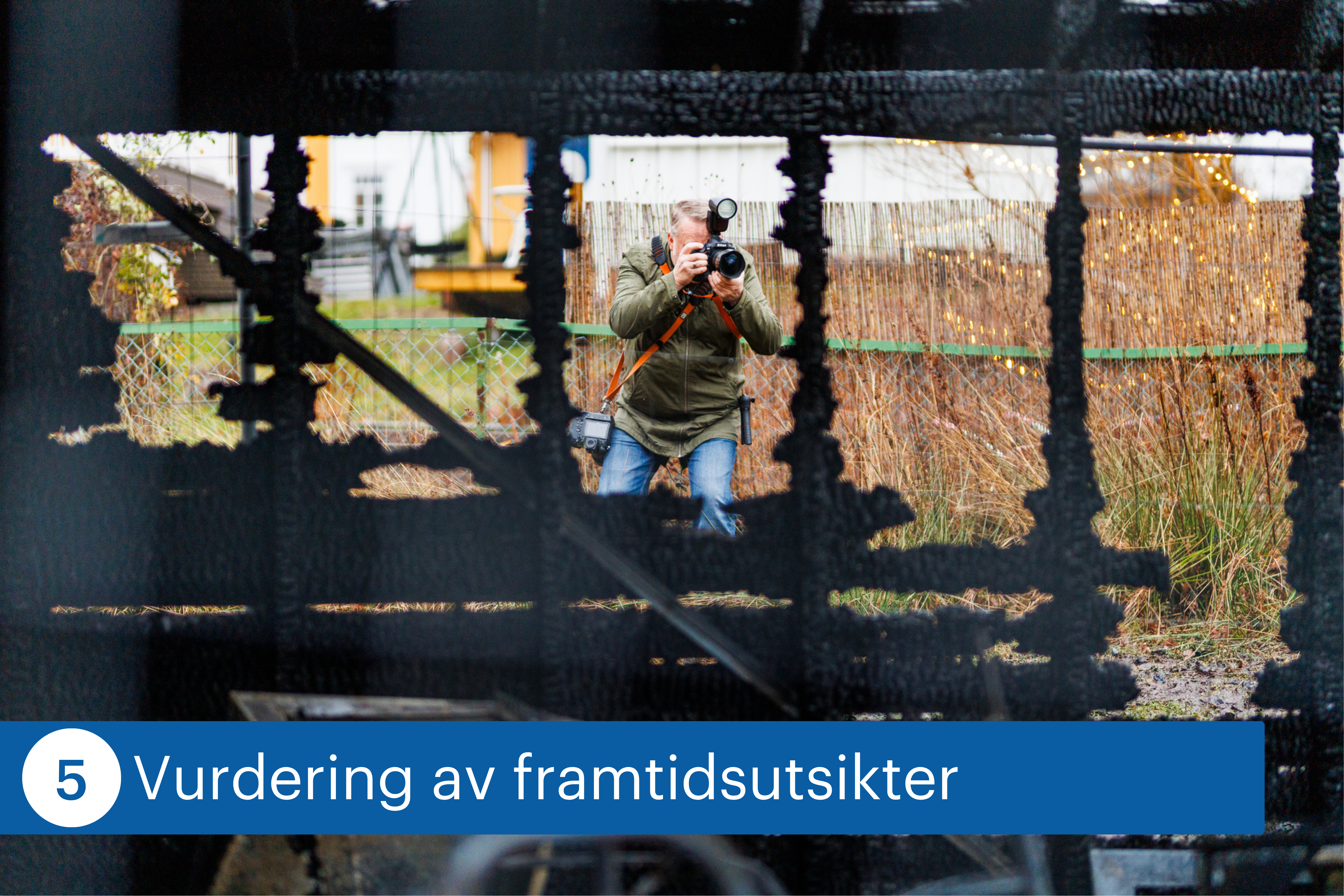 En fotograf tar bilder gjennom restene av en nedbrent bygning. I forgrunnen sees forkullede konstruksjoner, mens fotografen st&aring;r utenfor og dokumenterer skadene.