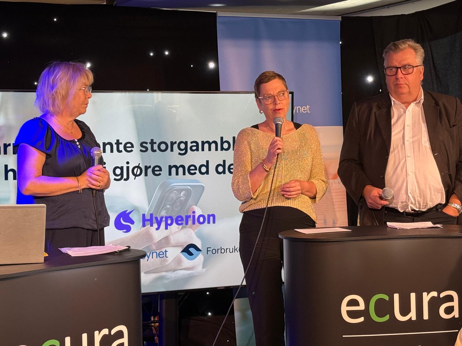 Tre personer st&aring;r p&aring; en scene med mikrofoner under et arrangement. I bakgrunnen vises logoene til Hyperion og Forbrukerr&aring;det p&aring; en skjerm.
