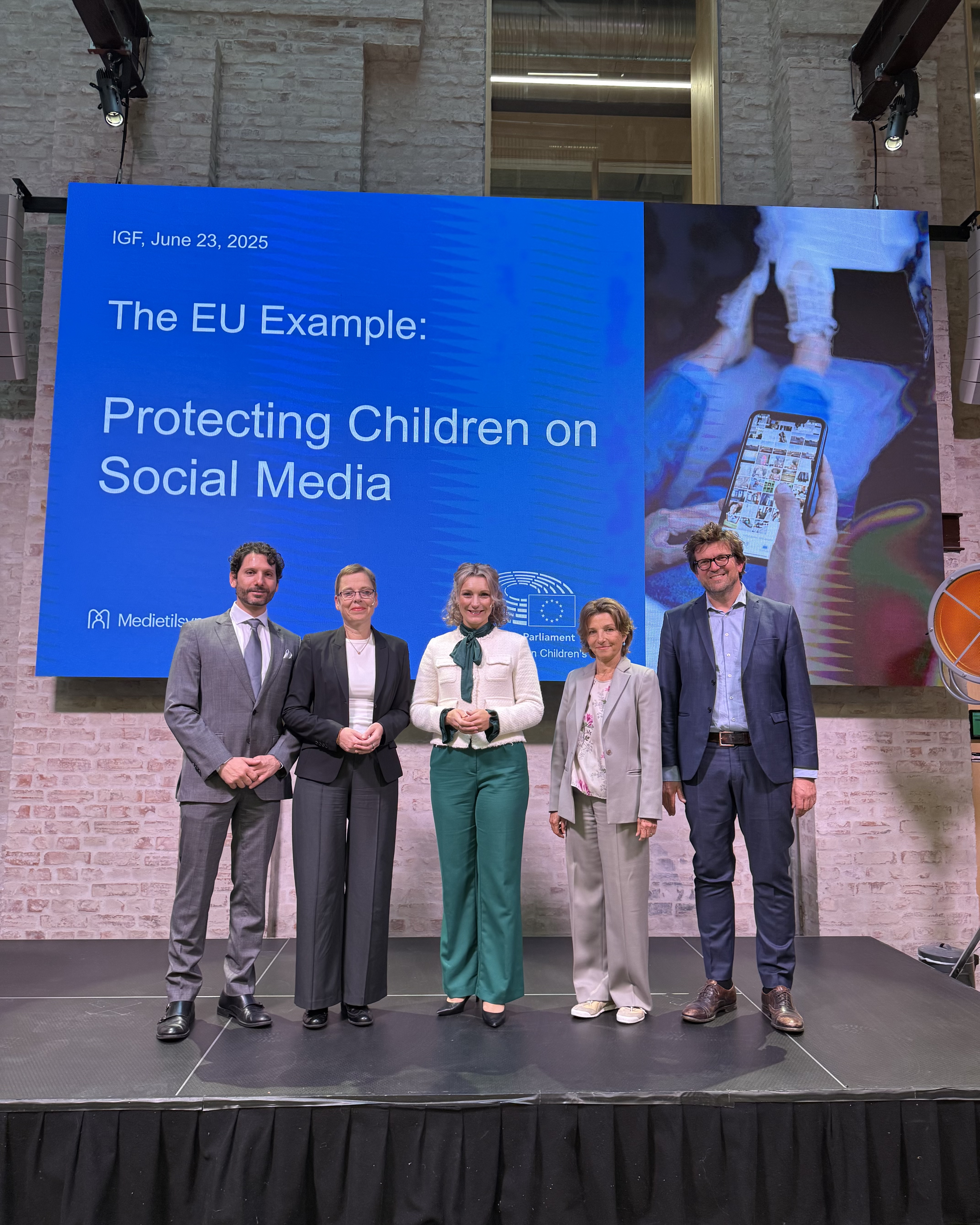 Fem personer st&aring;r p&aring; en scene foran en stor skjerm med teksten &laquo;The EU Example: Protecting Children on Social Media&raquo; under Internet Governance Forum 2025.