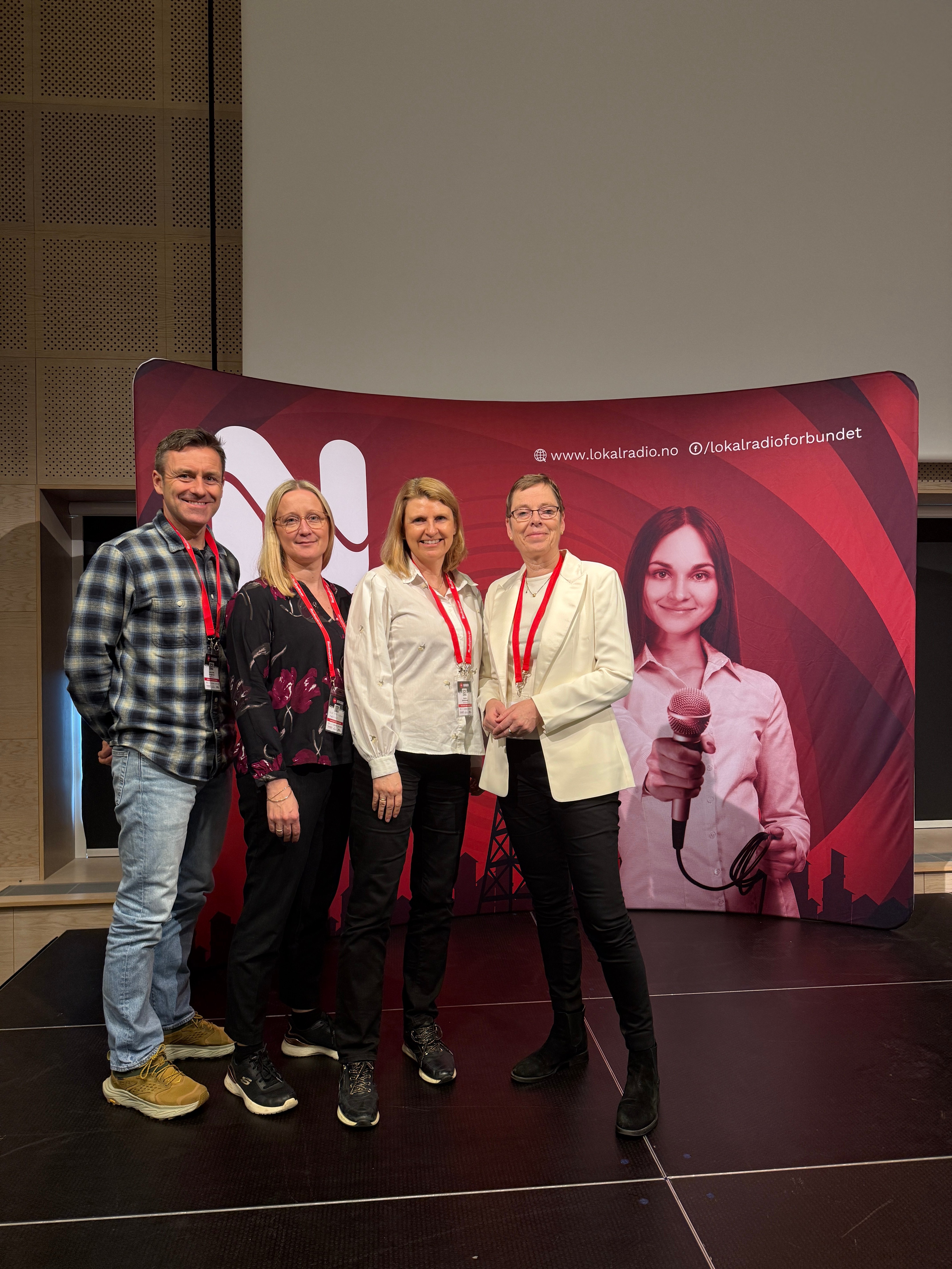 Fire representanter fra Medietilsynet st&aring;r foran en r&oslash;d messevegg med logo og nettadresse til Norsk lokalradioforbund under Lokalradiokonferansen.