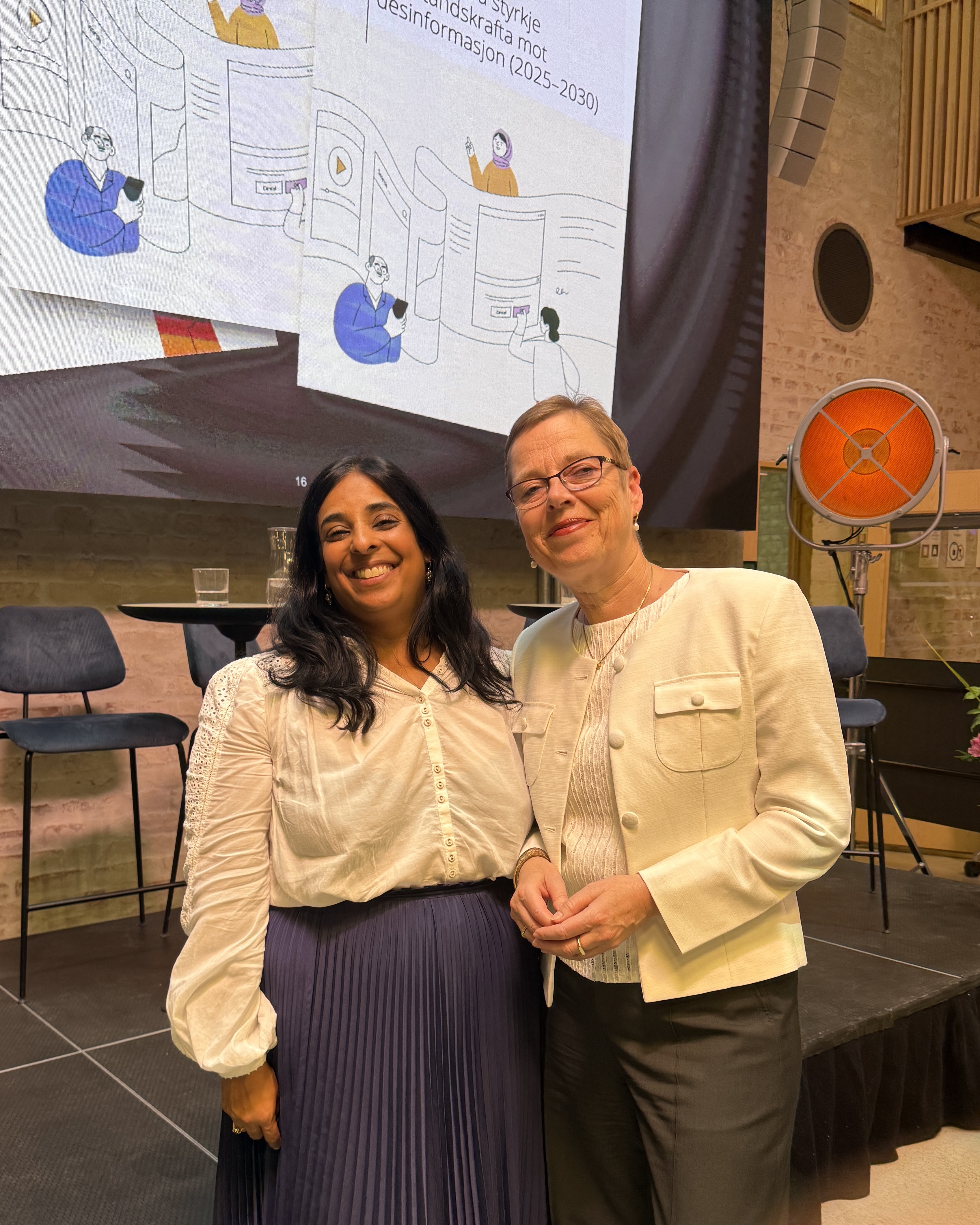 Lubna Jaffery og Mari Velsand st&aring;r smilende foran en scene der regjeringens strategi for &aring; styrke motstandskraften mot desinformasjon vises p&aring; storskjerm.