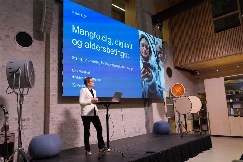 En person holder foredrag fra en scene med talerstol og PC. P&aring; en stor skjerm bak vises en presentasjon med tittelen &laquo;Mangfoldig, digitalt og aldersbetinget&raquo;, om status og utvikling i bruksmangfoldet i Norge.