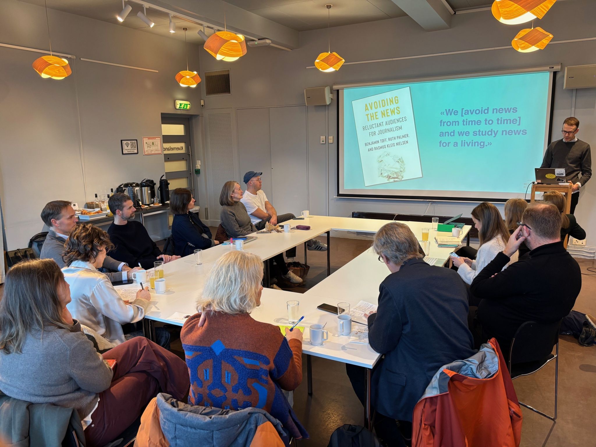 En person holder presentasjon i et m&oslash;terom, mens deltakere sitter rundt bord og f&oslash;lger med. P&aring; skjermen vises en presentasjon om nyhetsunng&aring;else med bokomslag og sitat.
