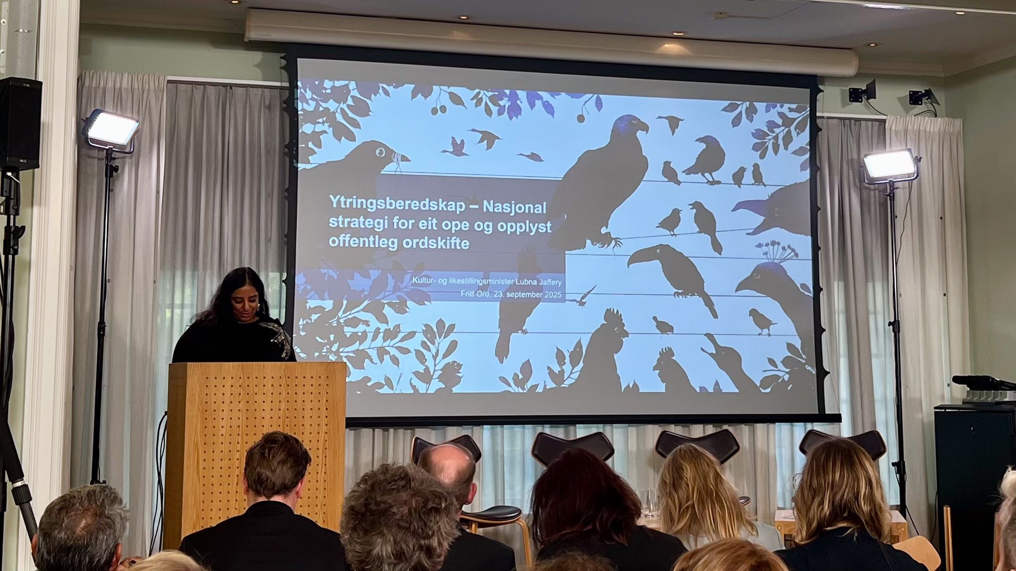 Foredrag med presentasjon av nasjonal strategi for ytringsberedskap. En person st&aring;r ved en talerstol foran en skjerm med illustrasjoner av fugler og tekst om offentlig ordskifte.