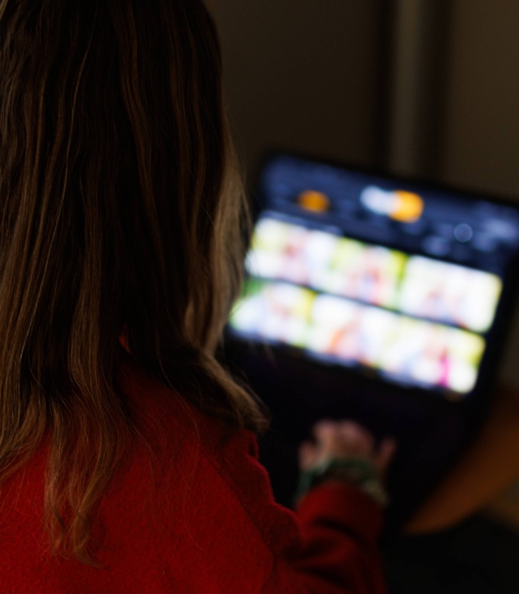 Person sitter i et m&oslash;rkt rom og bruker et nettbrett med flere bilder eller innholdsfelt p&aring; skjermen.